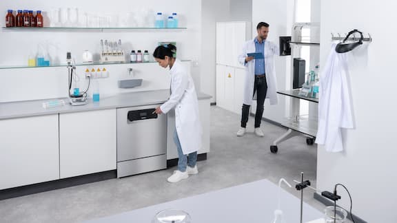 En laboratorietekniker betjener displayet på en laboratorieopvaskemaskine i rustfrit stål fra ExploreLine, mens en anden laboratorietekniker i baggrunden indtaster data på en skærm.