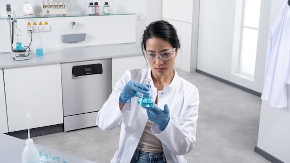 En laborant tester en blå væske i en lille erlenmeyerkolbe. I baggrunden ses en laboratorieopvaskemaskine i rustfrit stål fra ExploreLine i et rent og pænt laboratorium.