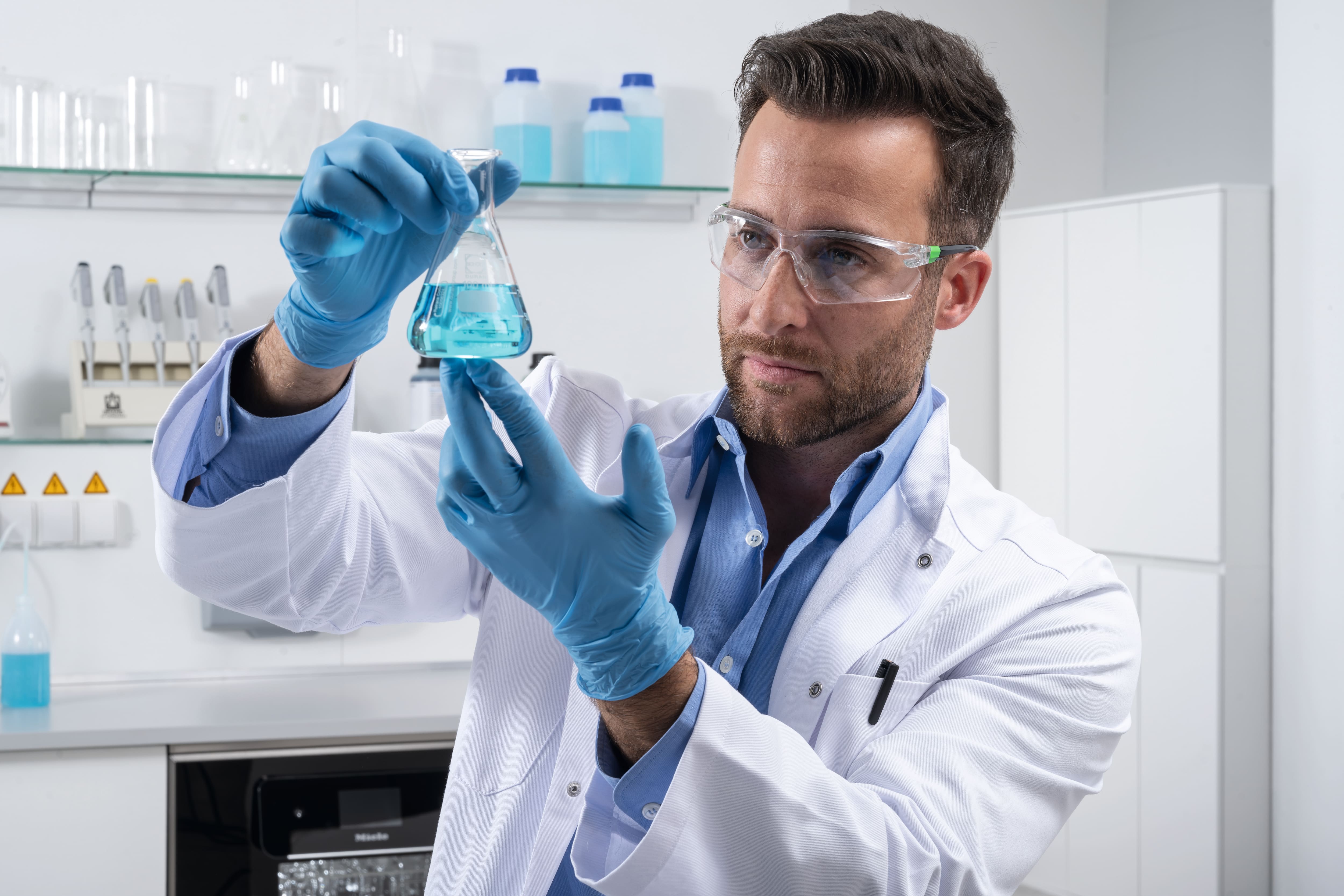 A laboratory assistant dressed in a white lab coat, gloves and safety glasses is holding an Erlenmeyer flask containing blue liquid.