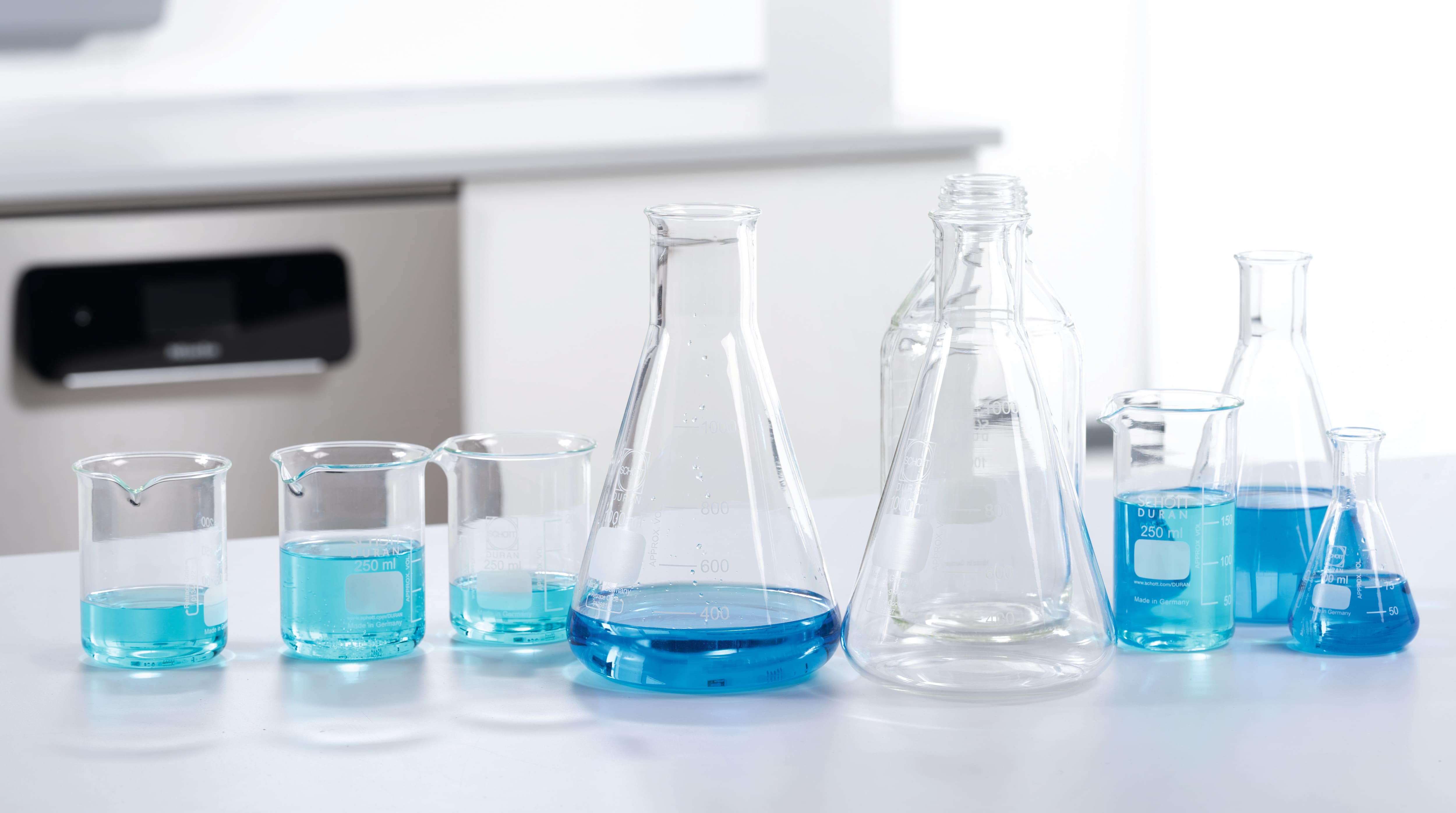 Verschiedene Laborgläser mit verschiedenen Flüßigkeiten stehen vor einer Untertischlaborspülmaschine auf einem Tisch.