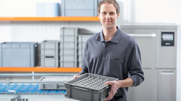 Ein Produktionsmitarbeiter steht vor einem Reinigungsgerät und hält eine Kiste mit Metall-Drehteilen in der Hand.