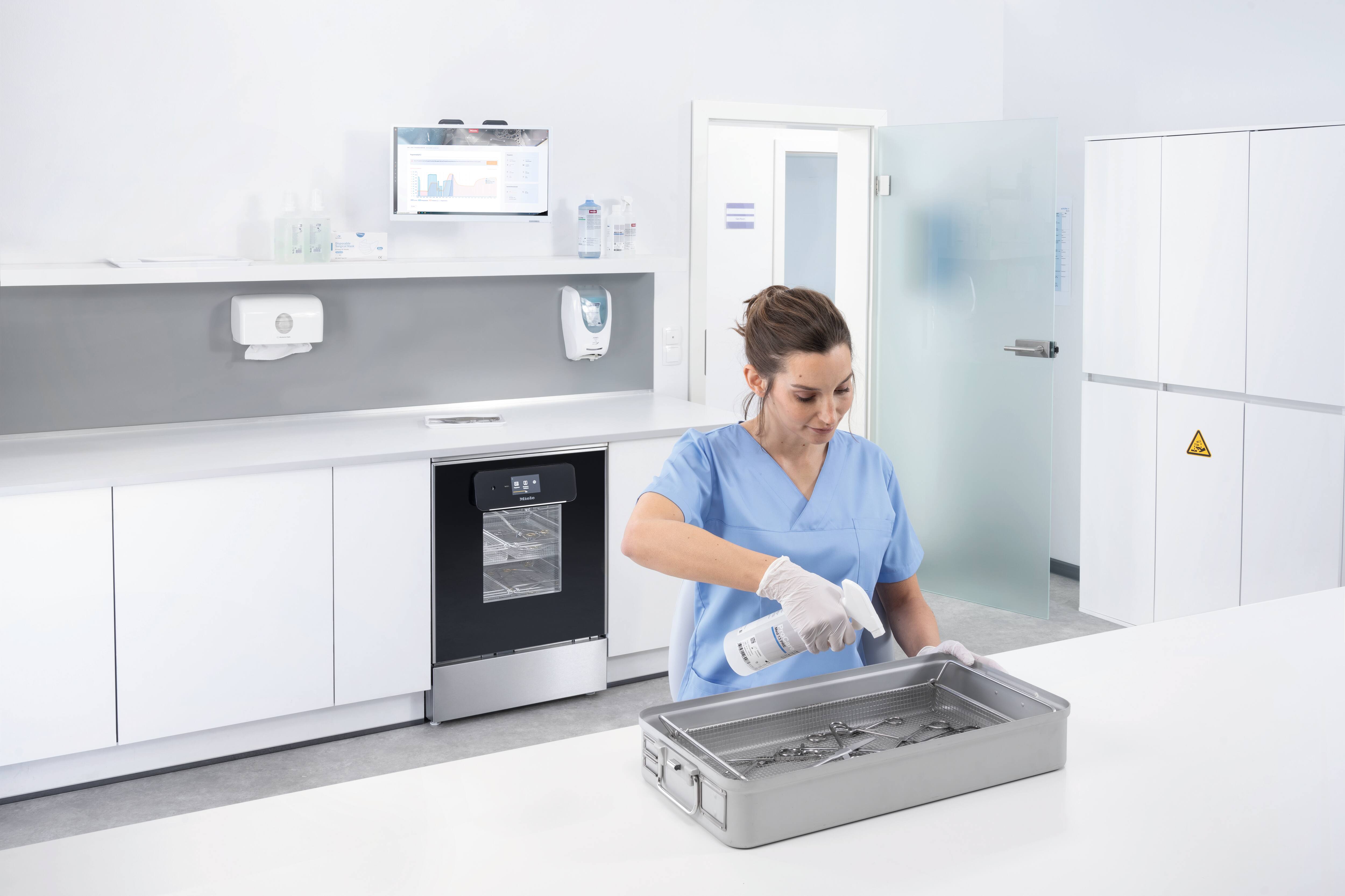 Eine medizinische Fachangestellte in einem Instrumentenaufbereitungsraum sprüht ProCare Med 13 PRE auf Instrumente zur Vorbehandlung, bevor diese gereinigt werden. Im Hintergrund ist ein Reinigungs- und Desinfektionsgerät PWD 8683 aus der ExpertLine-Serie von Miele Professional zu sehen.