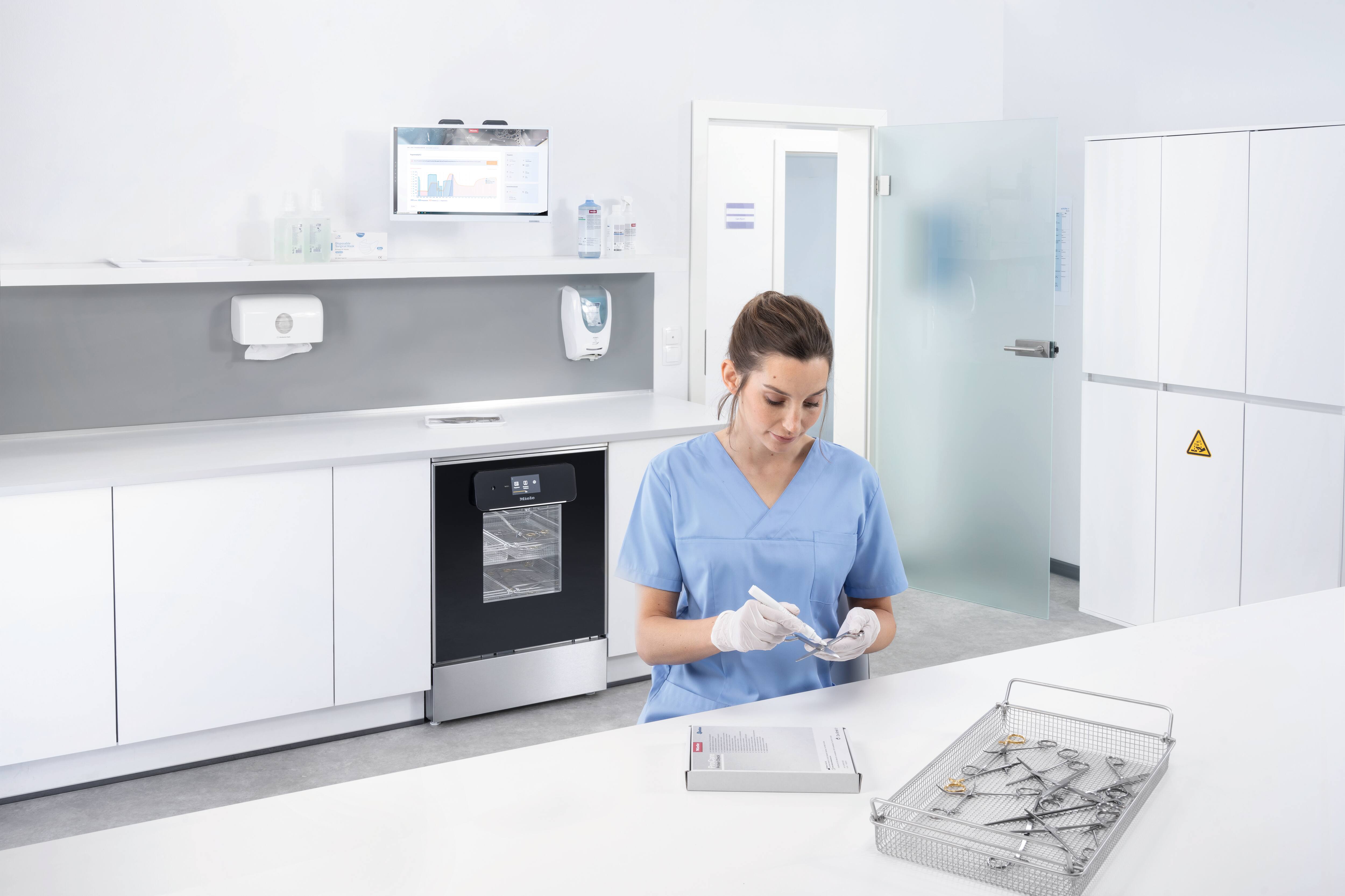 Eine medizinische Fachangestellte trägt ProCare Check Stick auf medizinische Instrumente in einem Netz-Tray auf. Im Aufbereitungsraum steht ein Miele Professional Reinigungs- und Desinfektionsgerät PWD 8682 in der Ausführung mit Glastür aus der ExpertLine-Serie. 