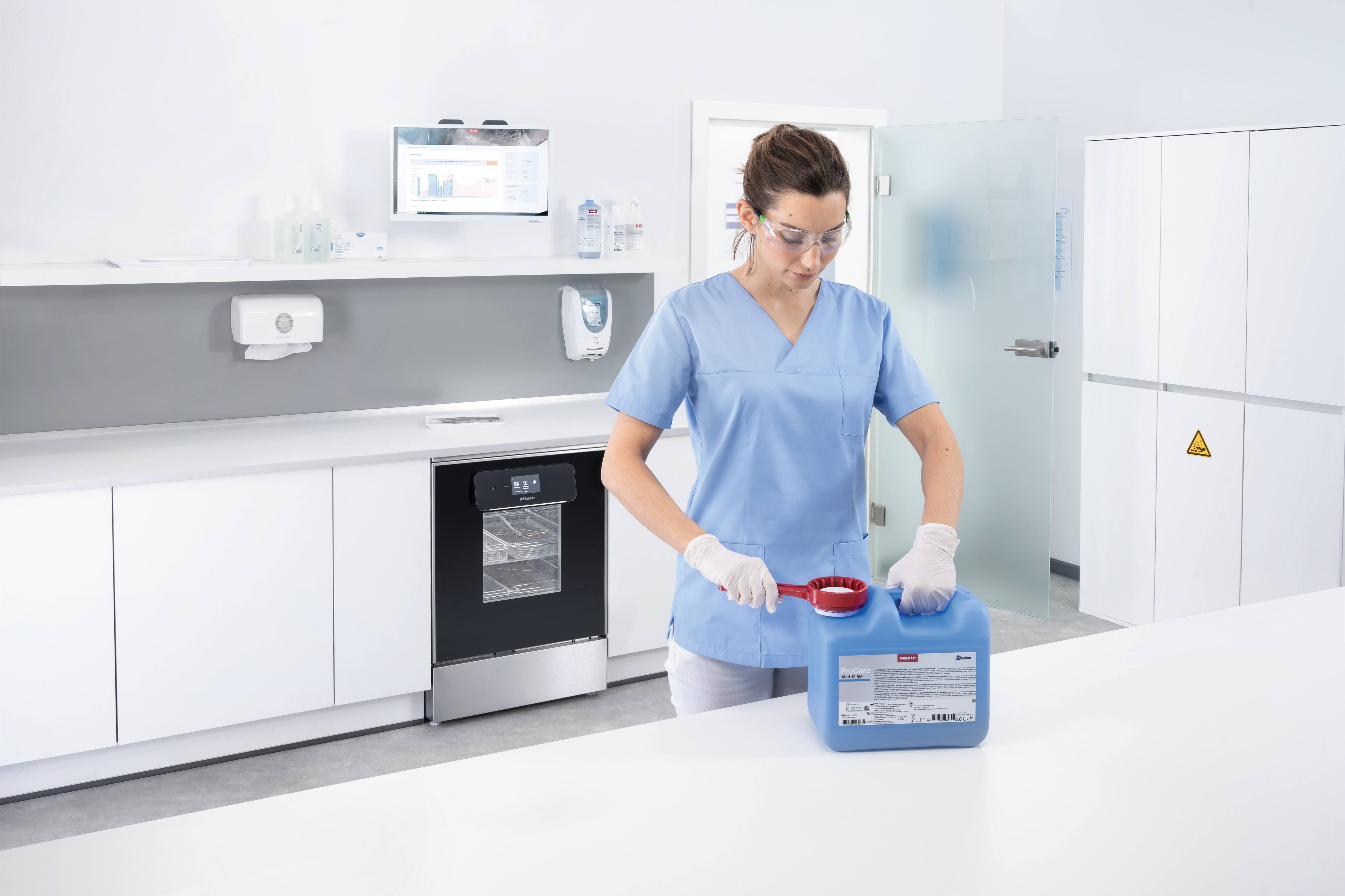 Eine medizinische Assistentin mit Schutzbrille öffnet einen Behälter mit ProCare Med-Spülmittel in einem Aufbereitungsraum mit dem Reinigungs- und Desinfektionsgerät Miele Professional ExpertLine PWD 8682.