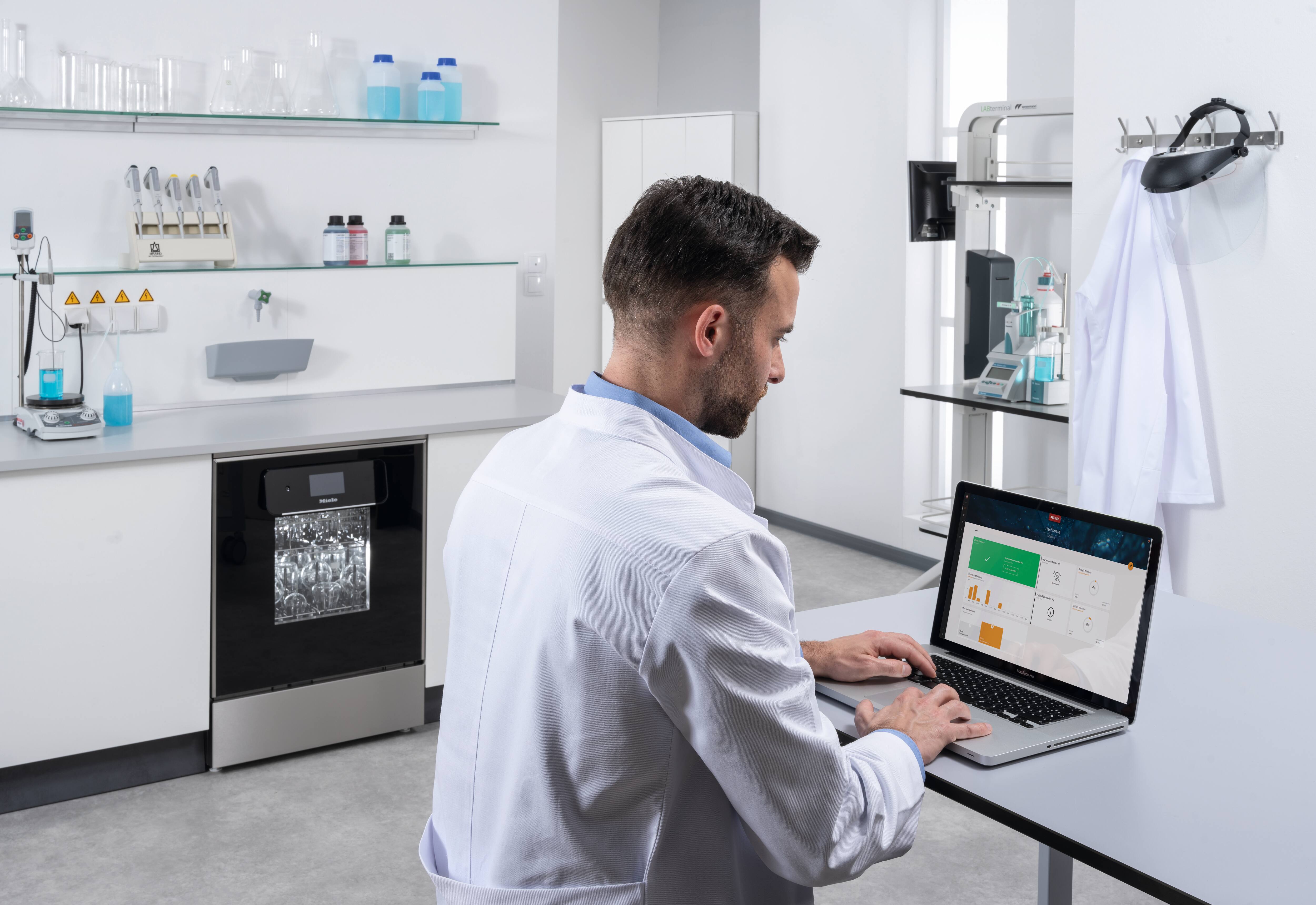 Ein Labormitarbeiter sitzt an einem Laptop, wo das Miele MOVE Connect Dashboard zu sehen ist. Im Hintergrund steht ein ExpertLine Laborspüler mit Glastür.