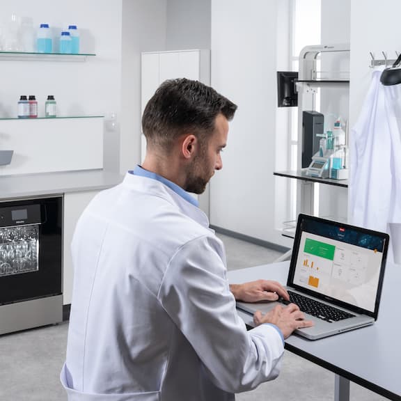 A laboratory assistant is sitting at a laptop where the Miele MOVE Connect dashboard can be seen. In the background is an ExpertLine laboratory washer with a glass door.
