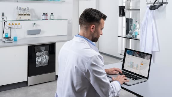 ラボ用ユニット(ExpertLineラボ用洗浄機を含む)が並ぶ前に、ノートパソコンを前にした検査技師がテーブルに座っている。ノートパソコンの画面にはMiele MOVE Connectが表示され、プログラムシーケンスが映し出されている。