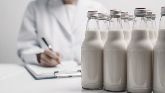 Kilka butelek mleka umieszczonych na stole; technik laboratoryjny robi notatki.