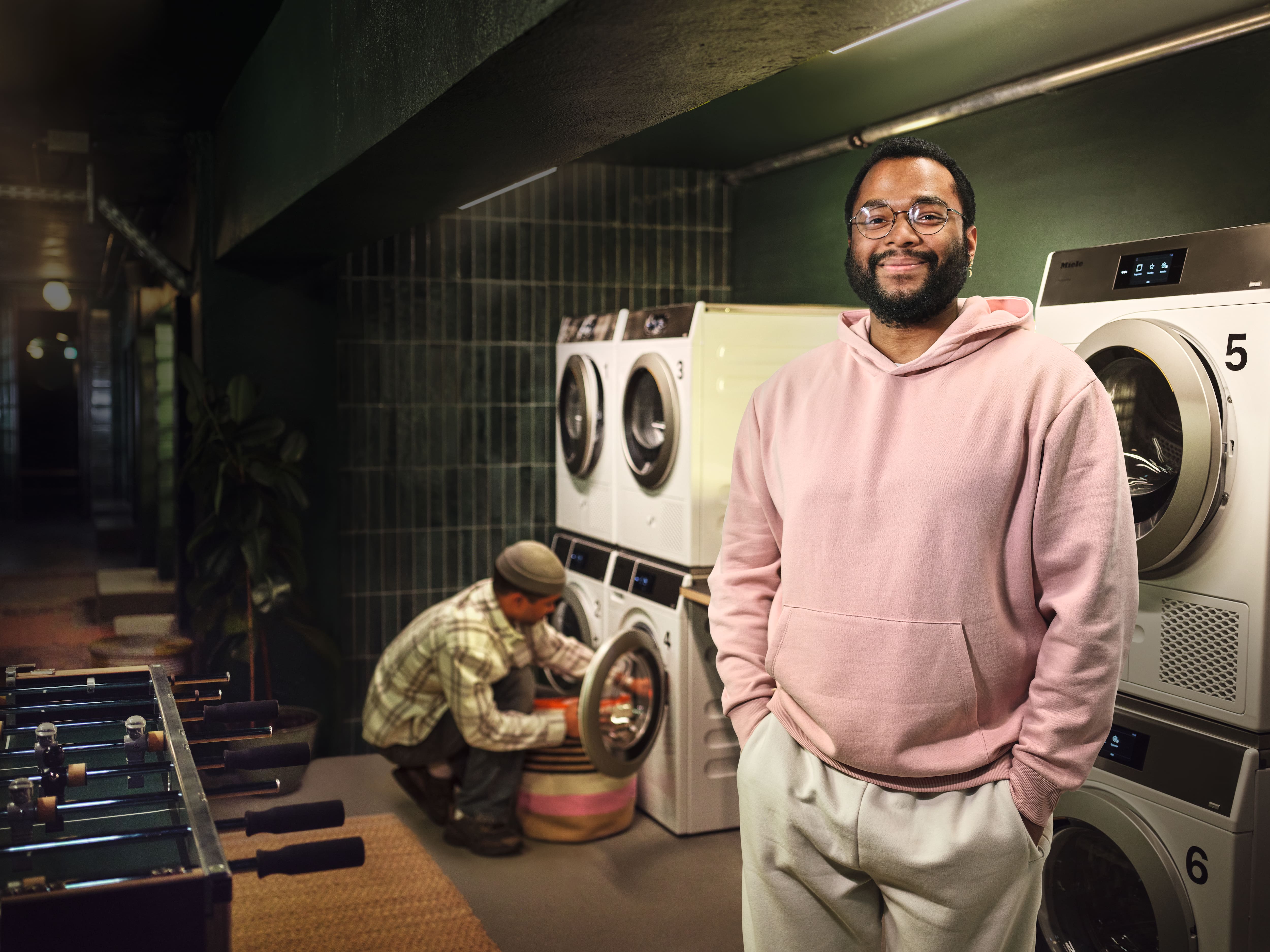 Ein junger Student steht in einem Waschkeller eines Studentenwohnheimes. Hinter ihm stehen die Kleinen Riesen Waschmaschinen und Trockner. Ein weiterer Student belädt im Hintergrund eine Waschmaschine.