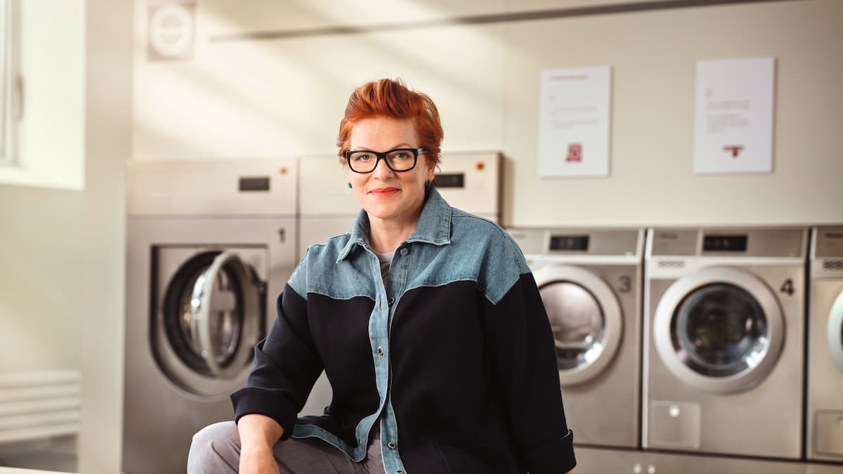 A middle-aged woman with glasses and a confident expression is shown in front of Miele PRO machines in a launderette