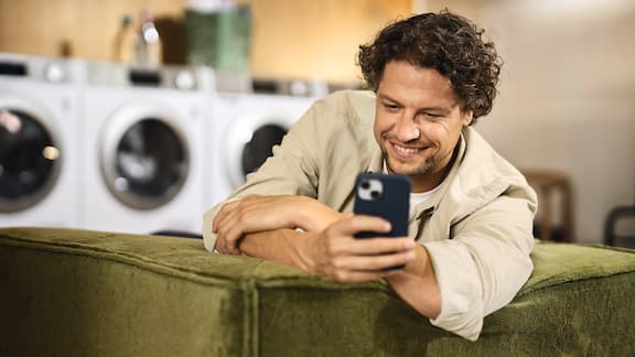 A man is sitting in a relaxed manner on a couch and is holding his smartphone in his hand. There are Little Giants washing machines and tumble dryers from Miele Professional in the background.