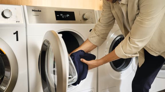 A person is unloading laundry from a lotus white Miele Professional Performance Little Giants tumble dryer.