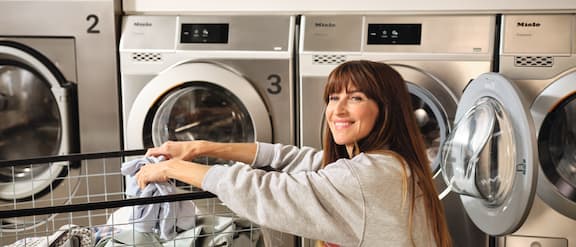 A woman squats in front of a Miele Professional Little Giants washing machine and rests her arms on a laundry trolley loaded with clothes.