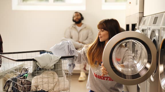 A lady squats in front of a Miele Little Giants washing machine and is loading her laundry into the machine. In the background, there is a young man with headphones who is waiting for his laundry.