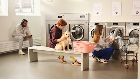 Two ladies are in a launderette. The lady on the left is sitting on a bench reading and stroking her dog while she waits for the laundry. The lady on the right is loading a Miele Professional Little Giants washing machine with laundry.