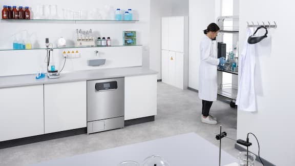 An ExpertLine PLW 8693 laboratory washer is installed in a laboratory run of units. In the background, a laboratory assistant is standing at a mobile workstation and dispensing liquid.