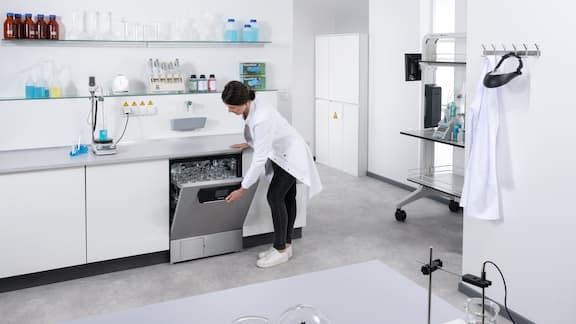 Eine Labormitarbeiterin öffnet einen Laborspüler mit einwandfrei getrocknetem Laborglas.