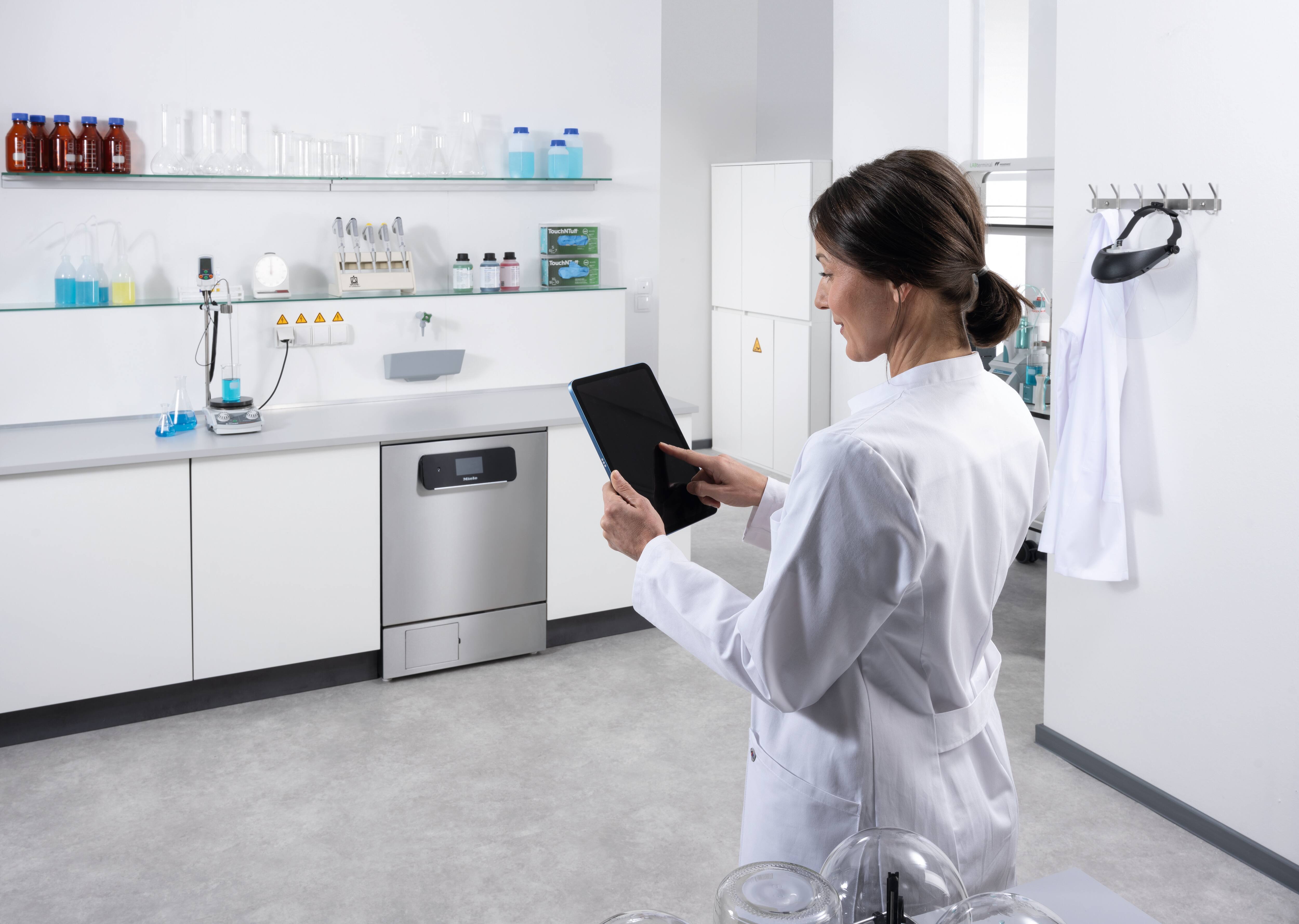 A lab technician sitting at a table with a laptop in front of a run of laboratory units including an ExpertLine laboratory washer. Miele MOVE Connect is displayed on the laptop screen with a programme sequence.