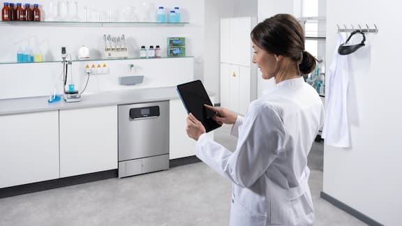 A lab technician sitting at a table with a laptop in front of a run of laboratory units including an ExpertLine laboratory washer. Miele MOVE Connect is displayed on the laptop screen with a programme sequence.