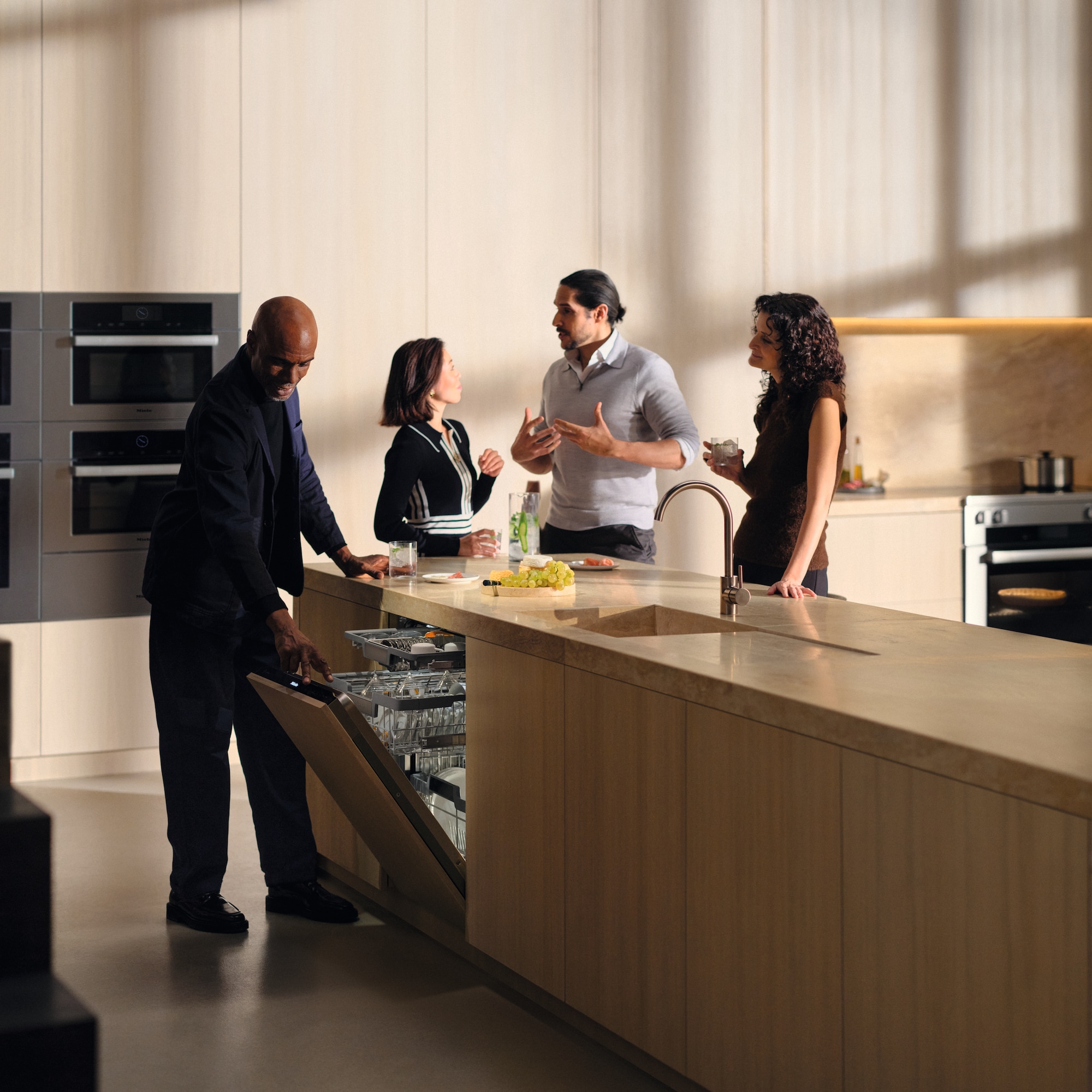 Group of friends talking around a kitchen island, while one of them opens a Miele Dishwasher to check it