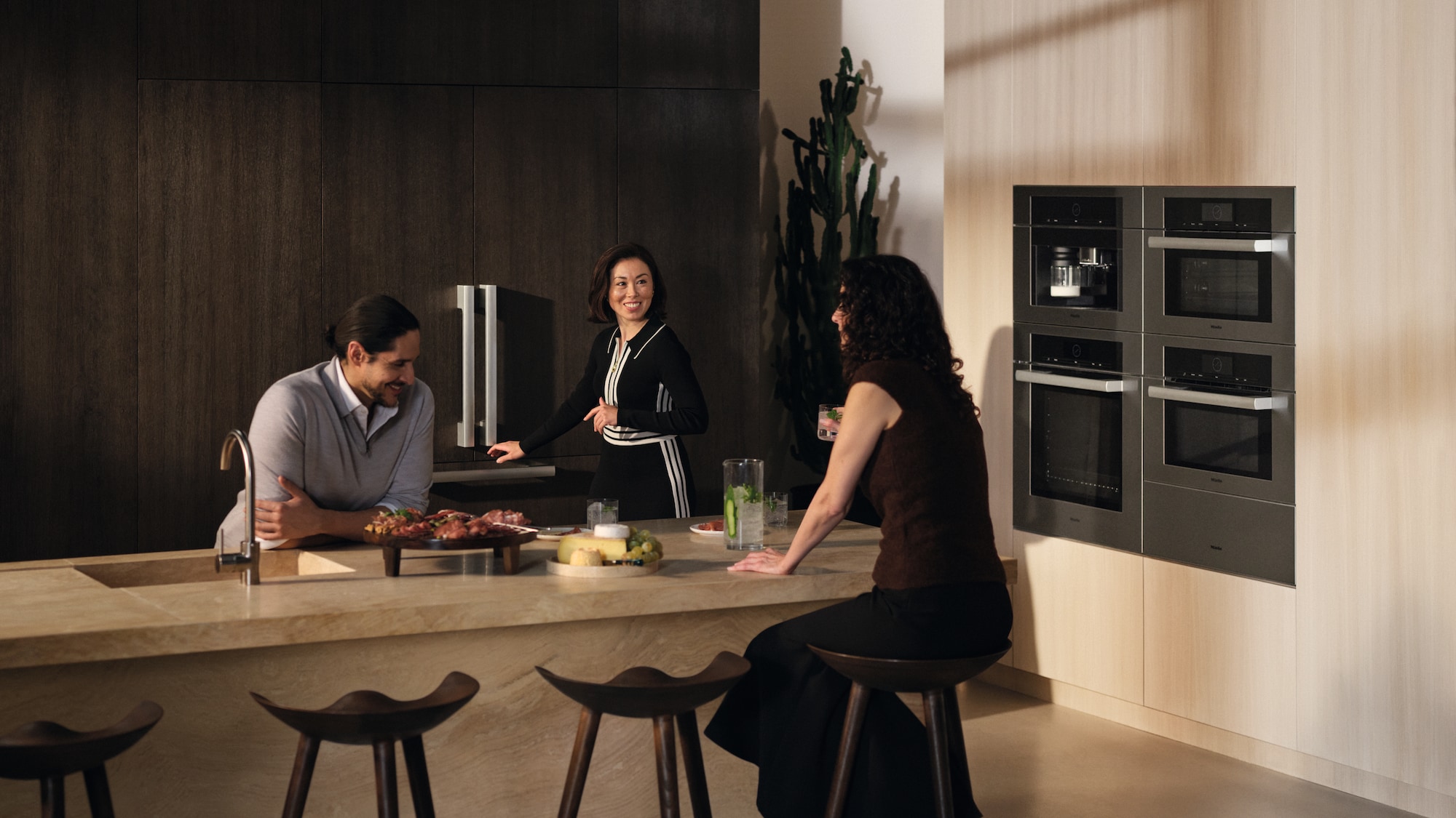 Three friends talking around a kitchen island while the host reaches to open the new Miele MasterCool refrigerator