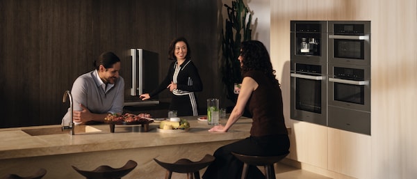 Three friends talking around a kitchen island while the host reaches to open the new Miele MasterCool Refrigerator