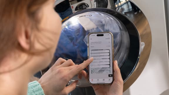 A young woman is holding a smartphone with the appWash app open in her hand. She is standing in front of a Miele Little Giants washing machine.