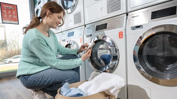 A young woman squats in front of a row of Miele Little Giants washing machines and looks at a smartphone. Next to her is a full laundry basket.