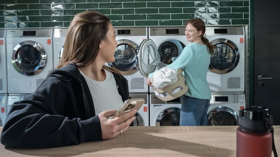 Two students are standing in a laundry room operated by appWash and chatting. The Miele Little Giants washing machines and tumble dryers can be seen in the background.