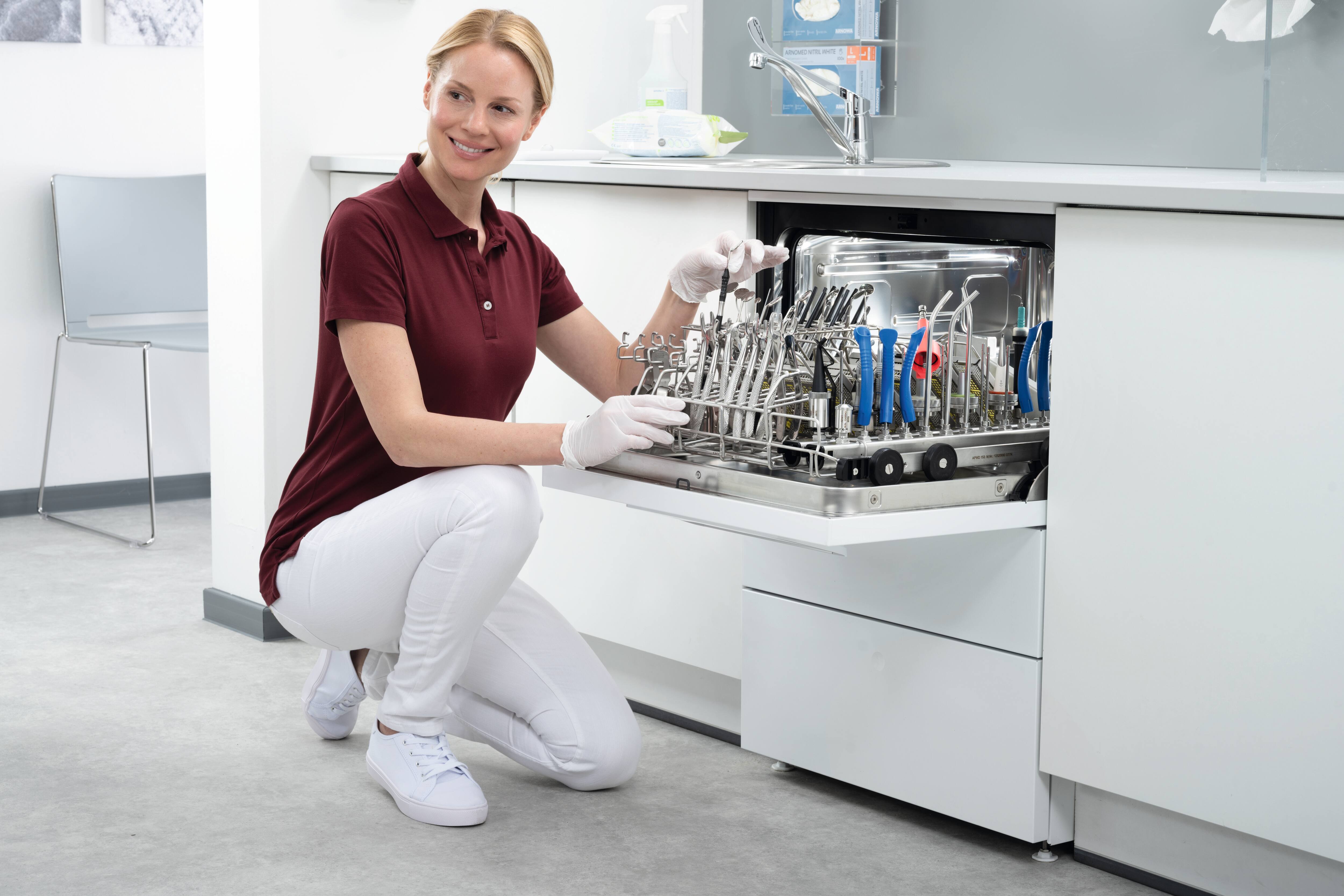 Eine Zahnarzthelferin lächelt vor einem mit zahnärztlichen Instrumenten beladenen CompactLine-Thermodesinfektionsgerät.