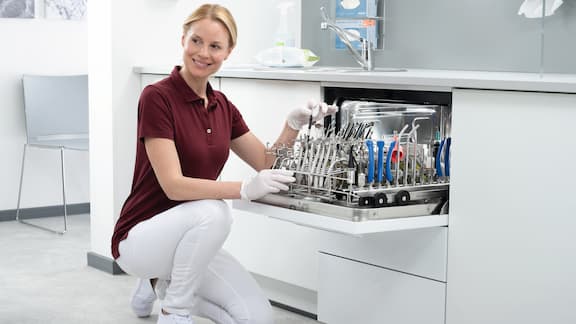 Une assistante agenouillée devant le laveur-désinfecteur CompactLine, porte ouverte, sourit.