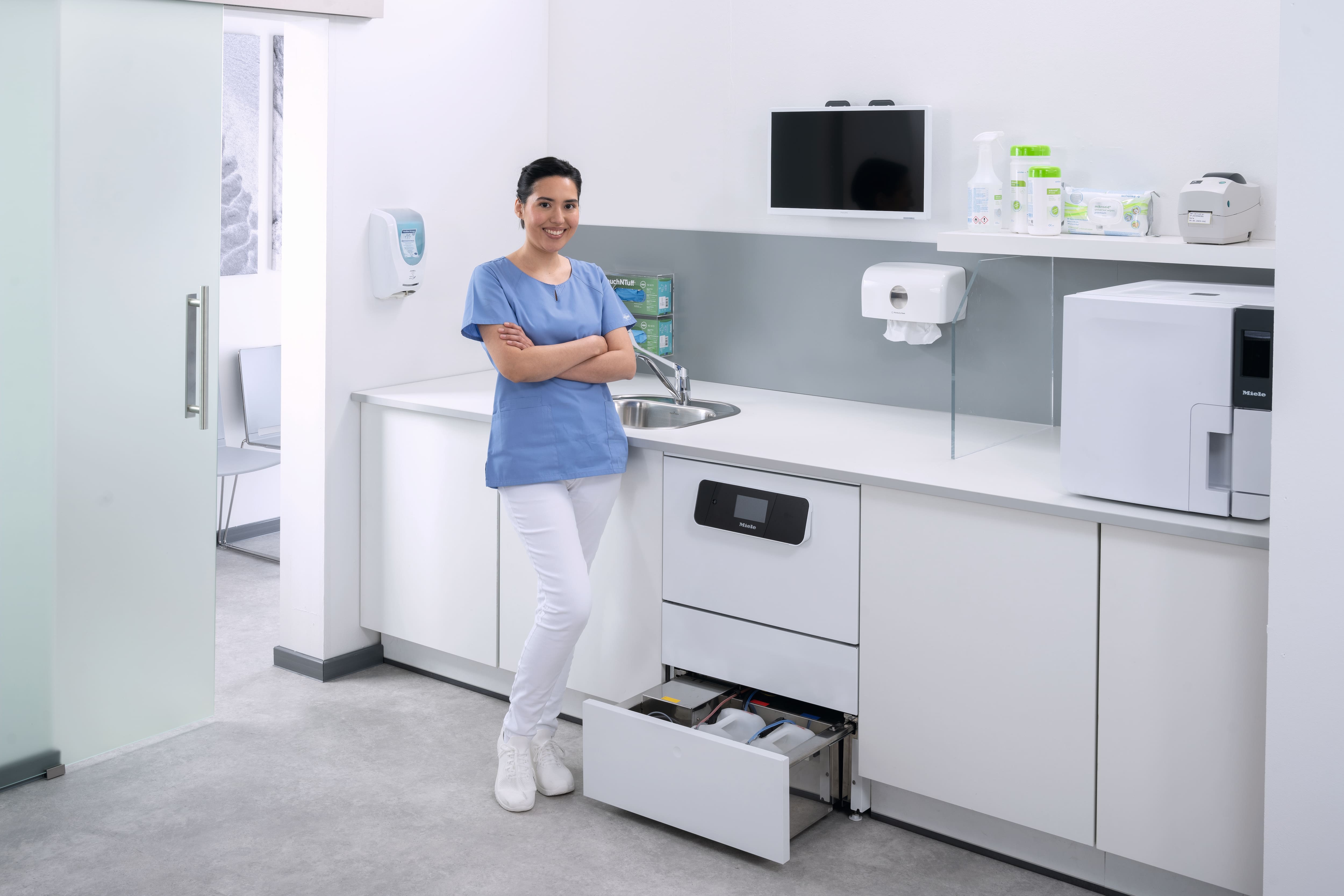 Eine medizinische Assistentin lächelt im Instrumentenaufbereitungsraum neben dem Reinigungs- und Desinfektionsgerät CompactLine mit offener Schublade.