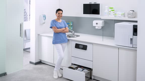 Uma assistente médica sorri nas salas de reprocessamento de instrumentos junto à máquina de lavar e desinfetar CompactLine com a gaveta inferior aberta