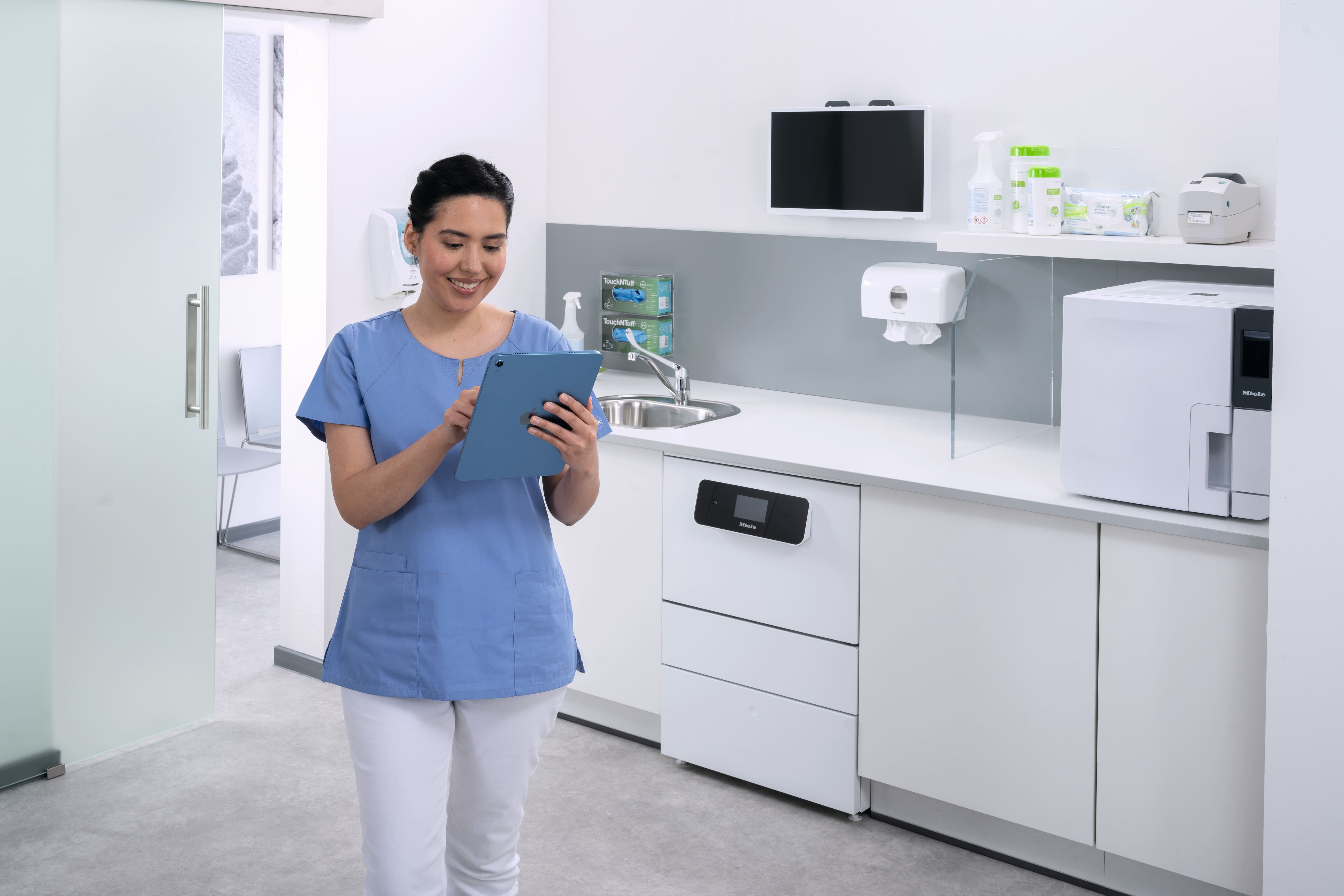 Eine medizinische Assistentin im Instrumentenaufbereitungsraum hält ein Tablet in der Hand. Im Hintergrund ist ein Reinigungs- und Desinfektionsgerät der CompactLine zu sehen.