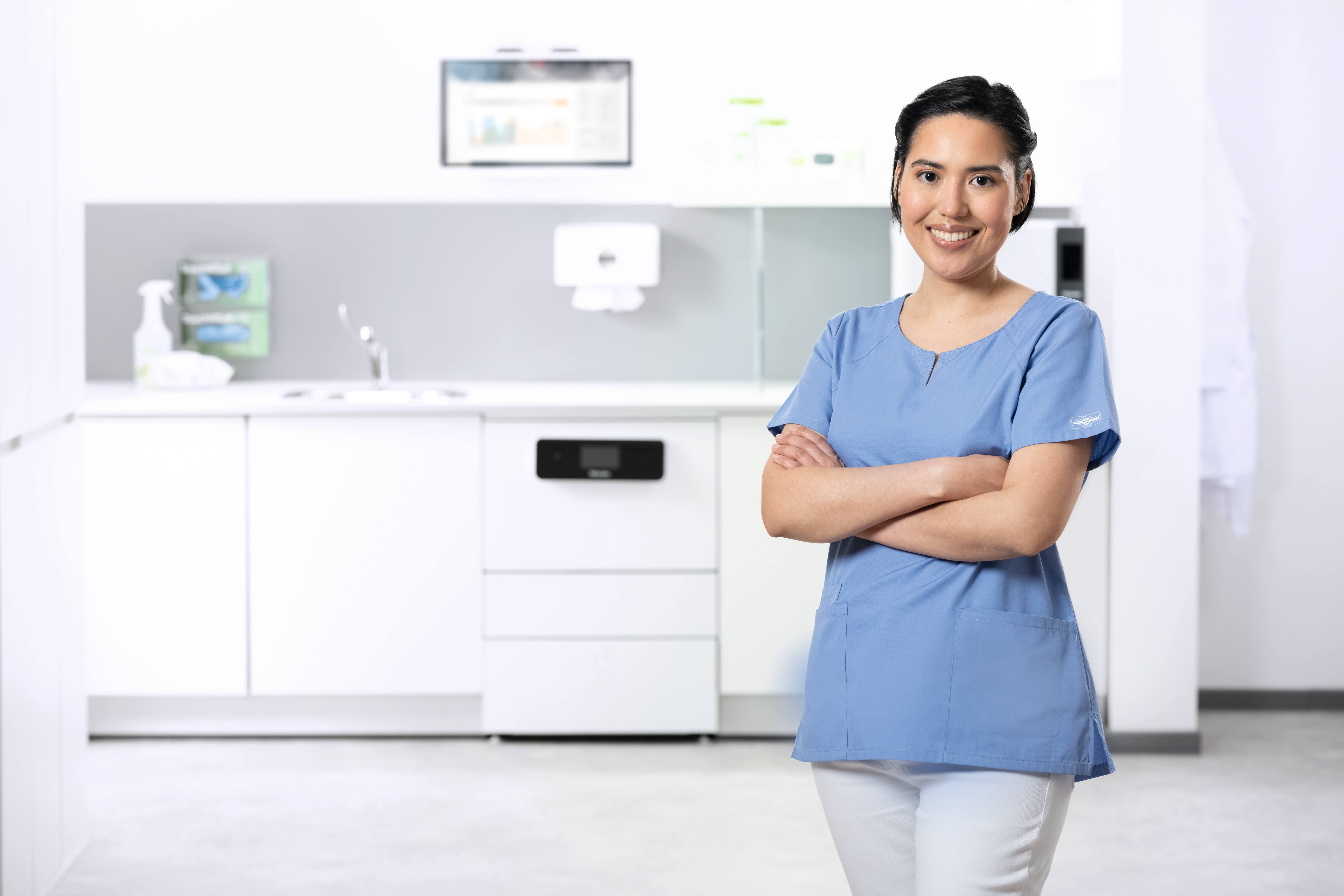 Eine medizinische Assistentin im Aufbereitungsraum lächelt in die Kamera. Im Hintergrund ist ein CompactLine-Reinigungs- und Desinfektionsgerät zu sehen.