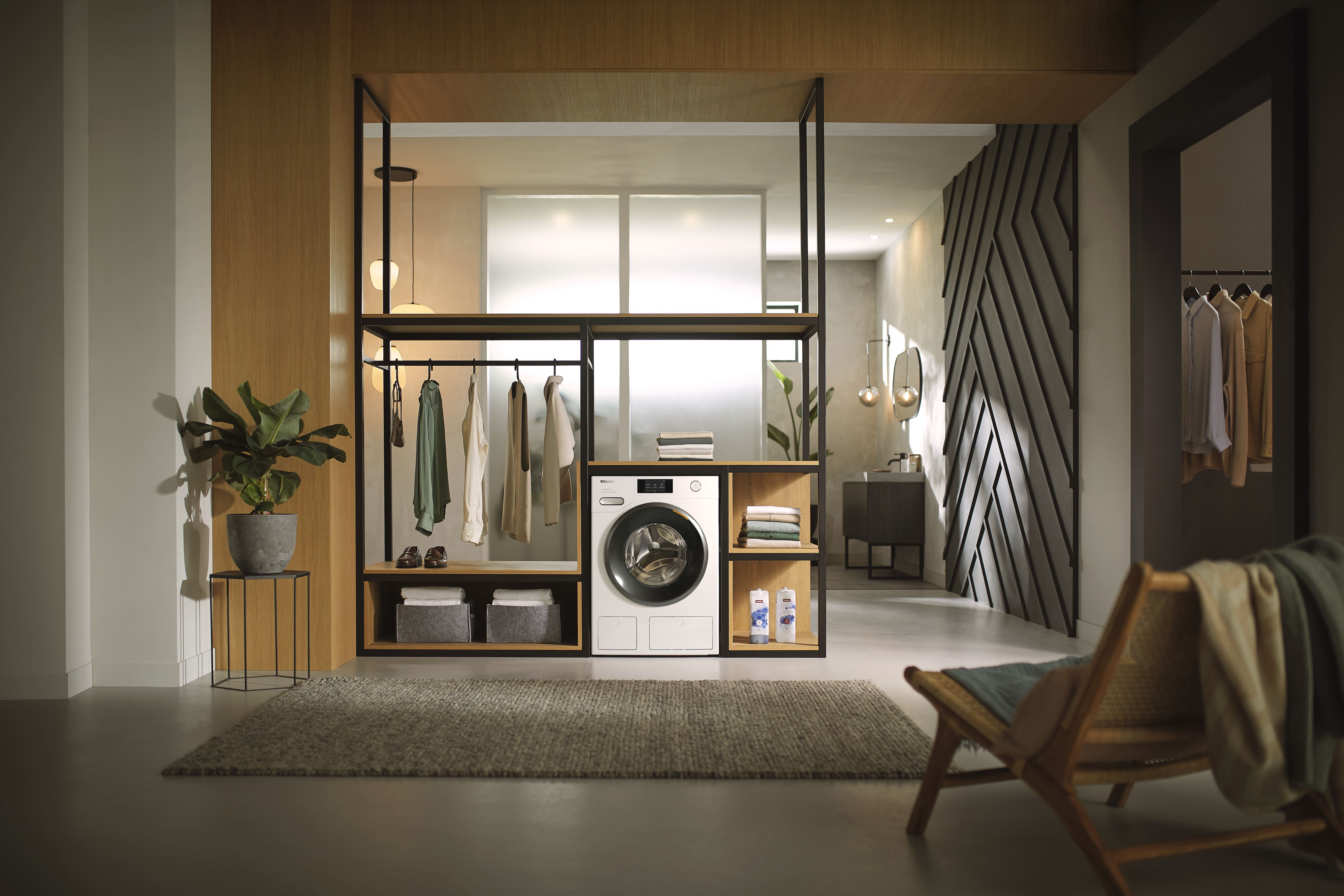 A modern open-concept laundry area with a washer integrated into a stylish shelving unit, surrounded by hanging clothes, folded linens, and storage compartments, set in a bright living space with plants and contemporary furniture. A modern open-concept laundry area with a washer integrated into a stylish shelving unit, surrounded by hanging clothes, folded linens, and storage compartments, set in a bright living space with plants and contemporary furniture.