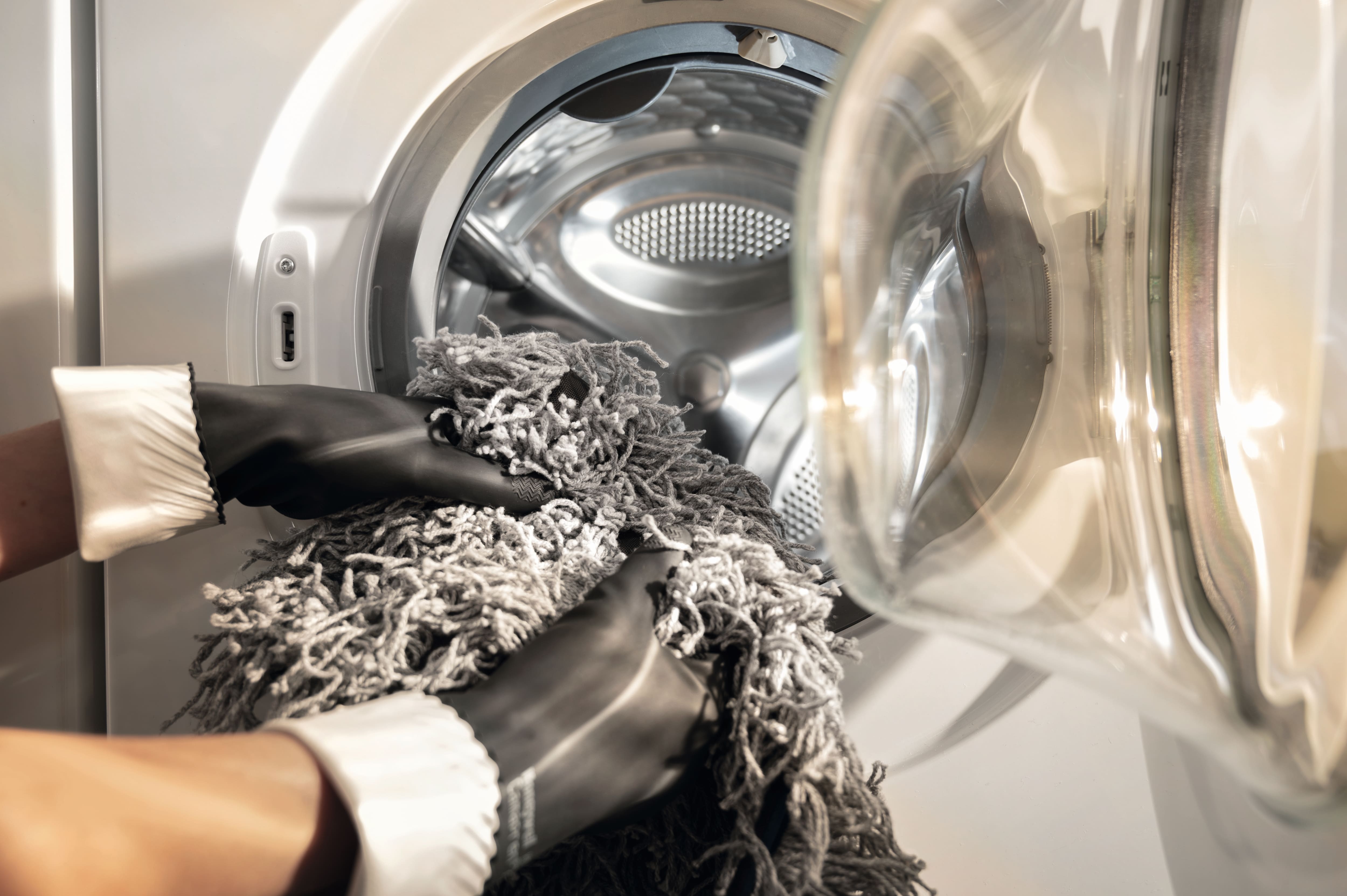 Person mit Handschuhen legt graue Mop-Textilien in die Trommel einer professionellen Miele Waschmaschine.