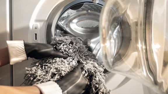 A person wearing gloves loads grey mop heads into the drum of a professional Miele washing machine.