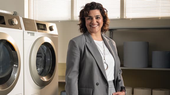 A person dressed in a business suit stands next to Miele Little Giants machines in a professional laundry setting.