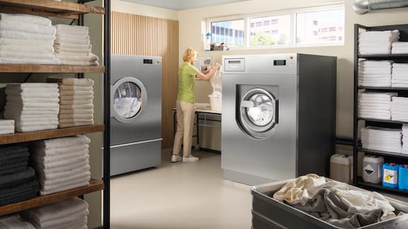 A person operates a commercial washing machine in a tidy, well-lit laundry room.