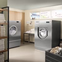 In a laundry room, there is a Benchmark Performance Plus PDR 928 tumble dryer and a Benchmark Performance Plus PWM 927 washing machine with a stainless steel look, as well as a table and industrial shelves filled with towels and ProCare Tex cleaning agents and care products.
