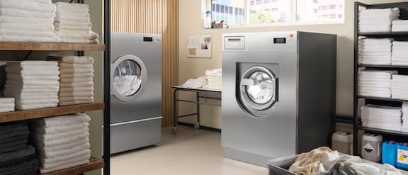 In a laundry room, there is a Benchmark Performance Plus PDR 928 tumble dryer and a Benchmark Performance Plus PWM 927 washing machine with a stainless steel look, as well as a table and industrial shelves filled with towels and ProCare Tex cleaning agents and care products.