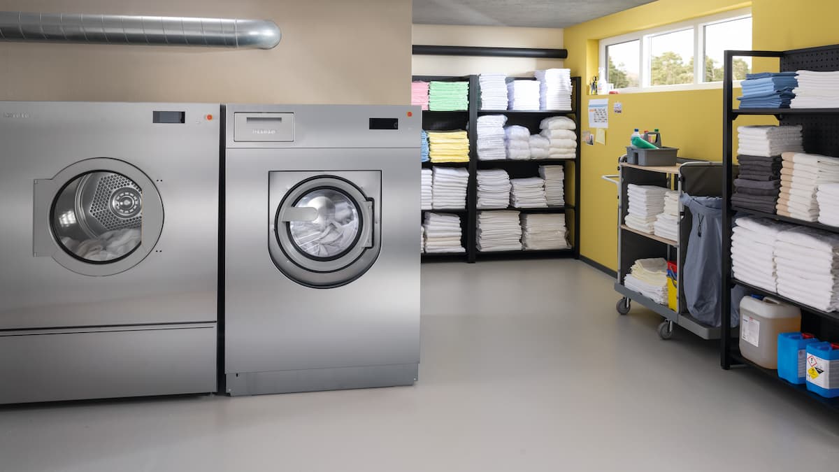 A laundry room with two Miele Professional machines – dryer on the left, washing machine on the right. The space is tidy, with colorful towels, cleaning supplies, and storage units against yellow walls