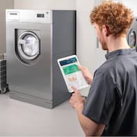A carer is standing with her back to the camera in an on-premise laundry. The carer is looking at a tablet on which the Miele MOVE Connect application is running. The laundry is equipped with a Miele Benchmark PWM 927 washing machine and a PDR 928 tumble dryer.