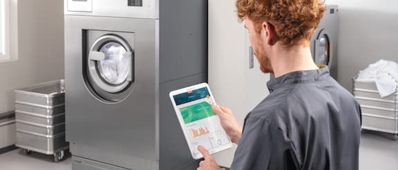 A carer is standing with her back to the camera in an on-premise laundry. The carer is looking at a tablet on which the Miele MOVE Connect application is running. The laundry is equipped with a Miele Benchmark PWM 927 washing machine and a PDR 928 tumble dryer.