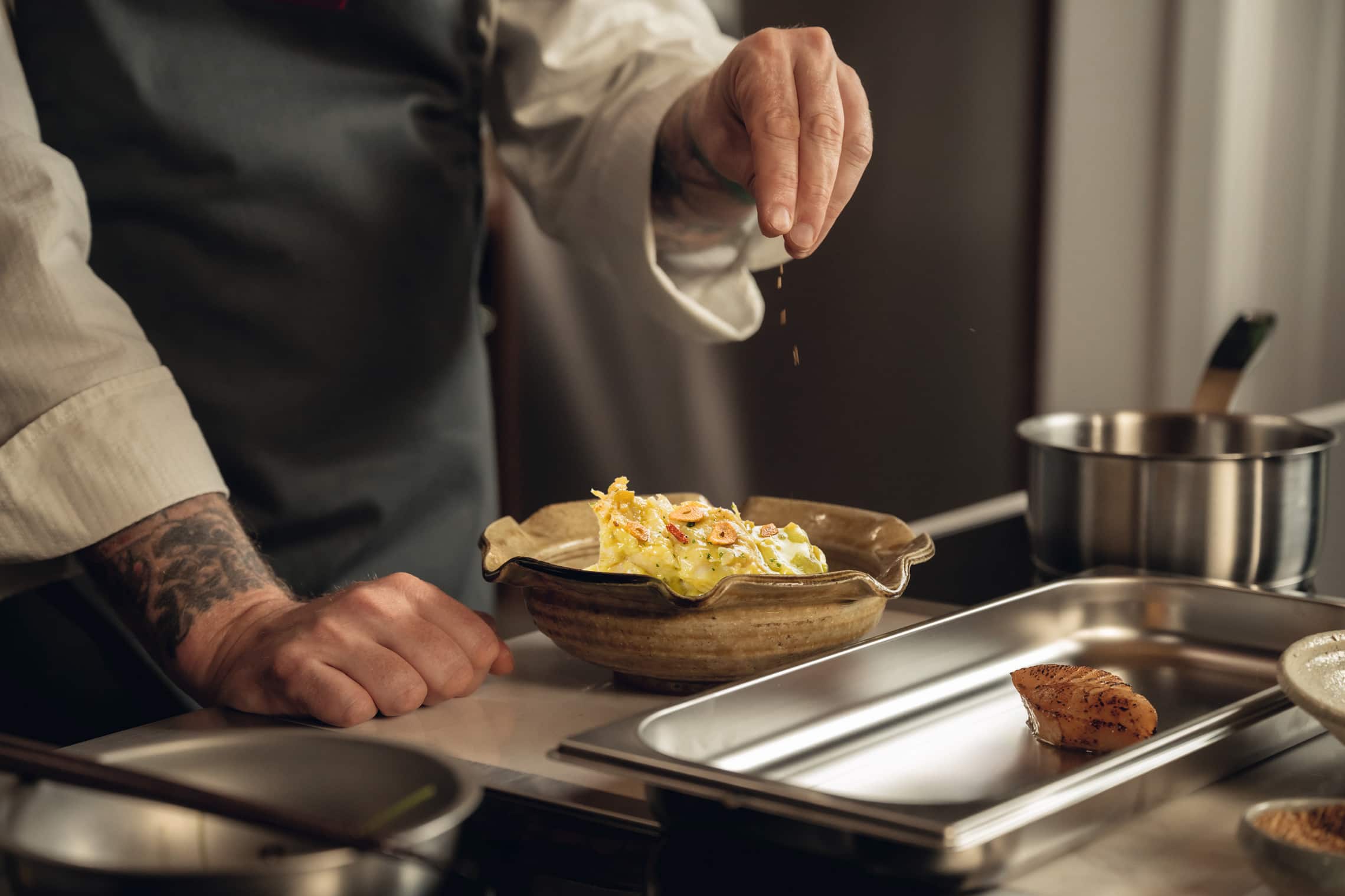 Chef sazonando un plato en una cocina, con utensilios y ingredientes visibles en el fondo