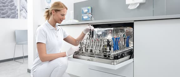 Een tandartsassistente knielt voor de CompactLine thermodesinfector met geopende deur en glimlacht