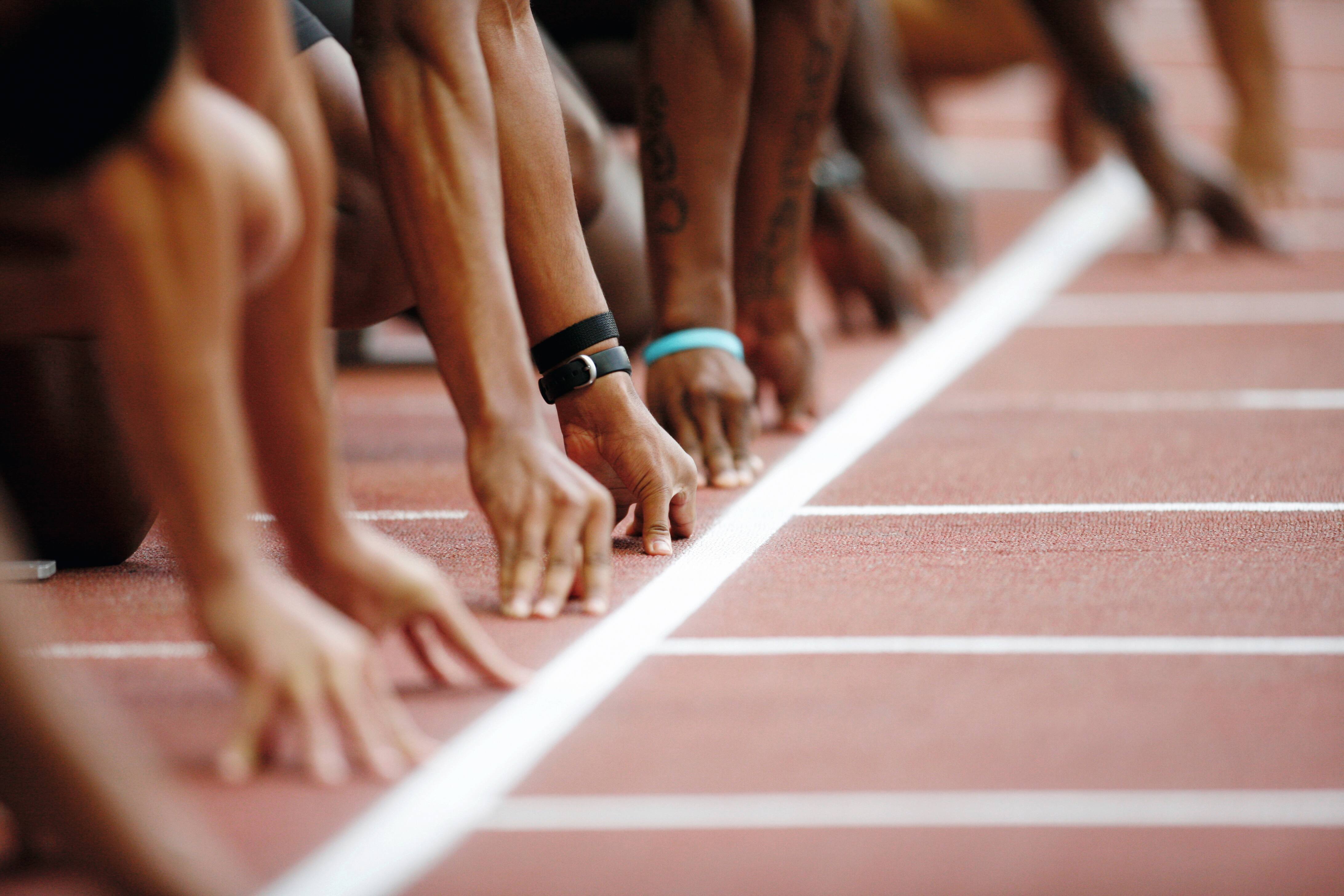 The sprinters’ hands are positioned side by side in front of the starting line for a race on a stadium track. 