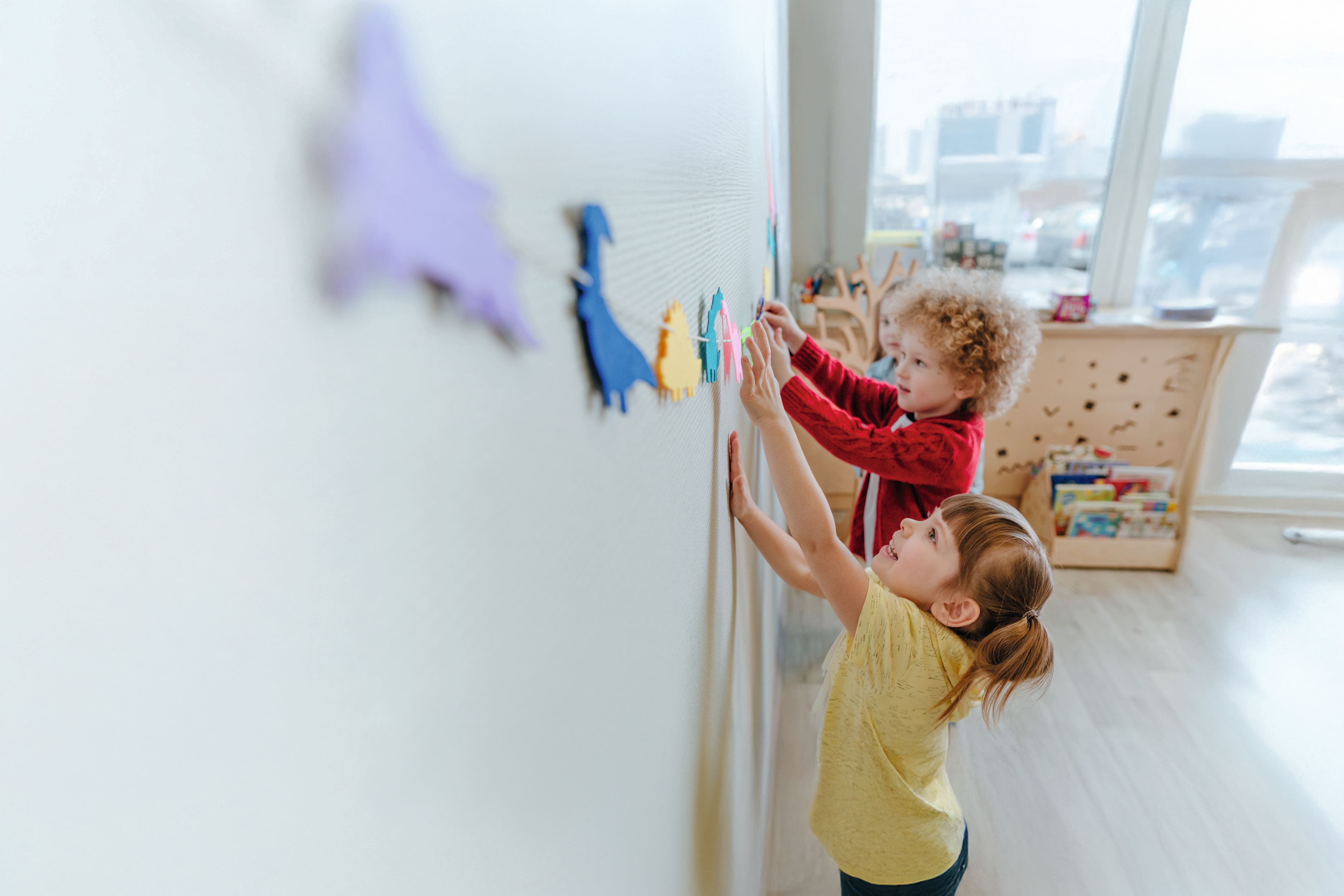 Zwei Kinder hängen bunte Papierfiguren an eine Schnur im Klassenraum und dekorieren gemeinsam die Wand.