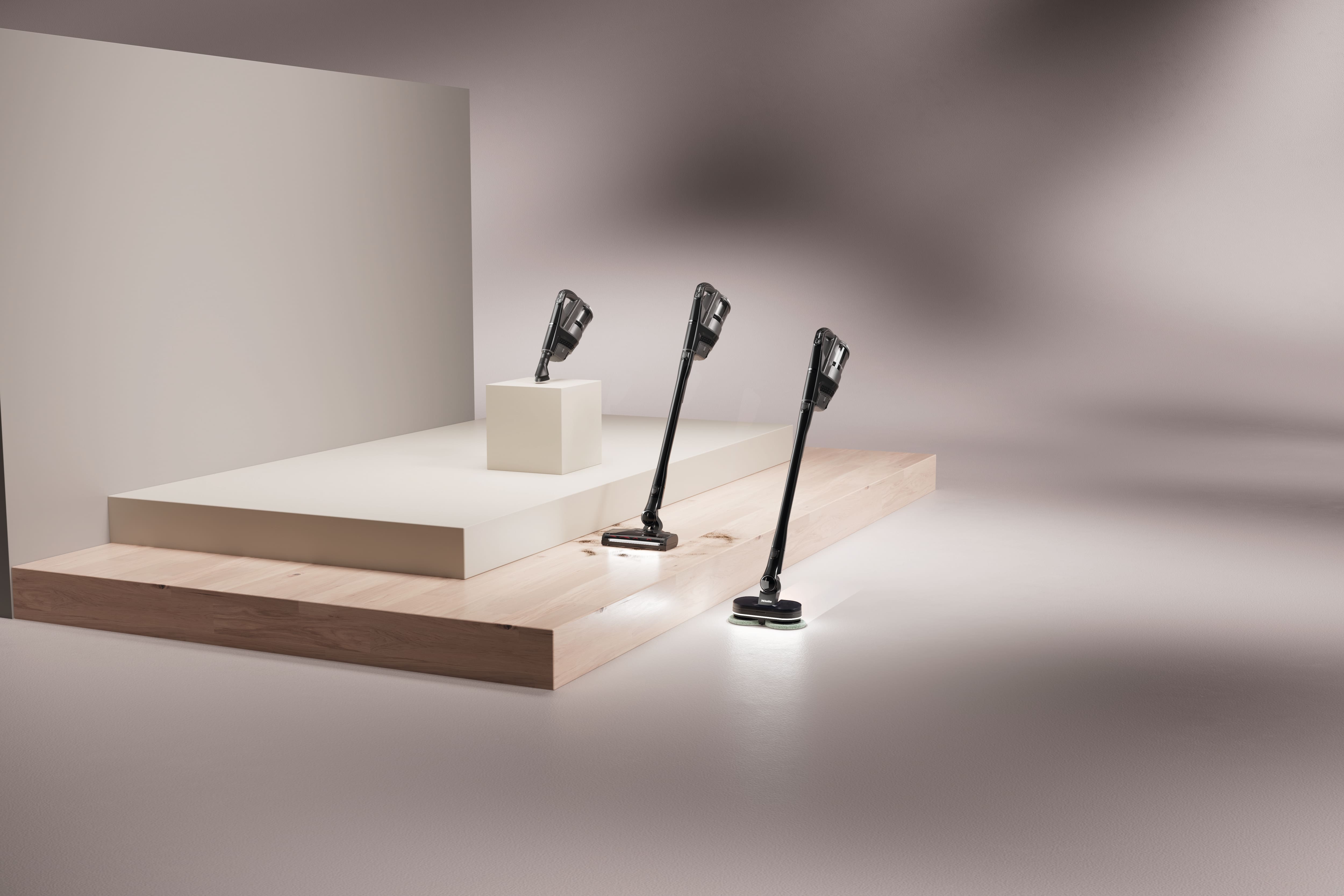 Tre aspirapolvere senza fili sono esposti su piedistalli minimalisti in uno studio moderno e luminoso con ombre morbide.